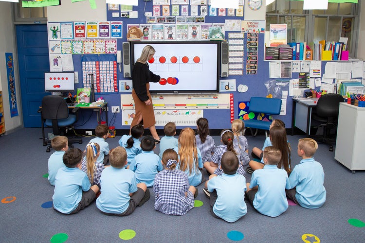 Students sitting on floor in class watching teacher at the whiteboard.